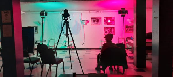 A room with fuchsia lighting, chairs and one person sitting in a chair in the foreground on the right 